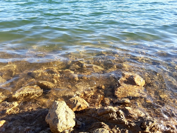 Clear water gently washes over a collection of stones and rocks, revealing their textures and colors beneath the surface. The sunlight creates a shimmering effect on the water, adding a sense of tranquility.