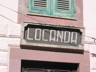 A weathered wooden sign with the word 'Locanda' is mounted on a rustic wall. Above the sign is a closed, old-fashioned, green-painted shutter window, and the wall surrounding the window and sign is constructed of rough, pinkish stone.