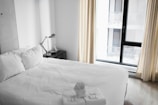 A neatly made bed with white linens is situated next to a modern-looking desk lamp on a dark nightstand. Large windows with light curtains allow natural light to enter the room, creating a bright and airy atmosphere. Towels are thoughtfully folded and placed on the bed, suggesting a welcoming and tidy space.