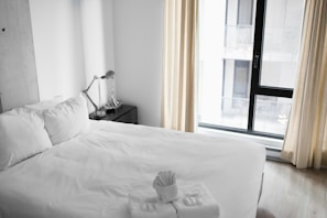 A neatly made bed with white linens is situated next to a modern-looking desk lamp on a dark nightstand. Large windows with light curtains allow natural light to enter the room, creating a bright and airy atmosphere. Towels are thoughtfully folded and placed on the bed, suggesting a welcoming and tidy space.