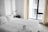 A neatly made bed with white linens is situated next to a modern-looking desk lamp on a dark nightstand. Large windows with light curtains allow natural light to enter the room, creating a bright and airy atmosphere. Towels are thoughtfully folded and placed on the bed, suggesting a welcoming and tidy space.
