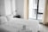 A neatly made bed with white linens is situated next to a modern-looking desk lamp on a dark nightstand. Large windows with light curtains allow natural light to enter the room, creating a bright and airy atmosphere. Towels are thoughtfully folded and placed on the bed, suggesting a welcoming and tidy space.