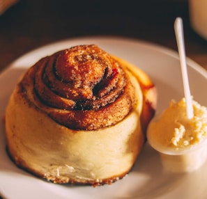A cinnamon roll topped with a drizzle of creamy icing, resting on a beige ceramic plate with a cinnamon stick beside it.