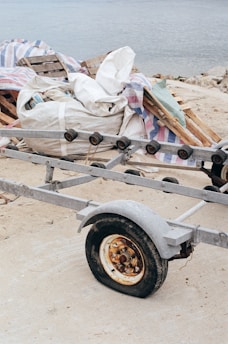 A rusted metal trailer with a single visible wheel is resting on a sandy surface. In the background, there are stacked wooden pallets and a large white bag, possibly filled with materials, draped with a colorful striped tarp. The scene overlooks a body of water.