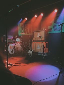 A dimly lit stage setup with musical equipment including a drum set, guitar amplifier, and electric guitar. Colorful stage lights in red, orange, blue, and purple create an atmospheric ambiance. Microphones and cables are scattered across the stage, indicating a live music performance setup.