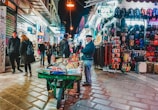 A bustling night market street scene with various stalls displaying an assortment of goods. People walk along the cobbled pavement, some browsing the products while others pass by. A vendor stands next to a cart filled with items, and the surrounding stalls showcase colorful backpacks, belts, and other merchandise. The area is illuminated by streetlights and shop signs.
