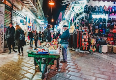 A bustling night market street scene with various stalls displaying an assortment of goods. People walk along the cobbled pavement, some browsing the products while others pass by. A vendor stands next to a cart filled with items, and the surrounding stalls showcase colorful backpacks, belts, and other merchandise. The area is illuminated by streetlights and shop signs.