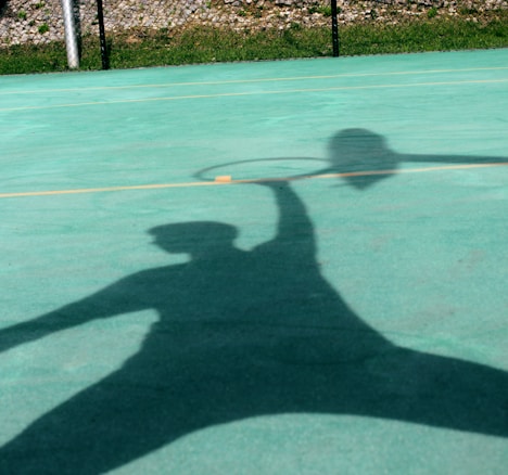 A large, distinct shadow of a person holding a tennis racquet is cast on an outdoor tennis court. The court surface is a bright aqua color, and the shadow indicates a dynamic pose, possibly capturing a moment of action, like a serve or a swing.