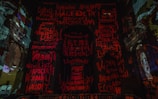 The image displays a dark, dramatic interior with walls covered in vibrant red and orange graffiti-style text. Architectural elements of what appears to be a historic building are visible. A portrait of a face is discernible on the right side.