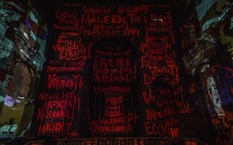 The image displays a dark, dramatic interior with walls covered in vibrant red and orange graffiti-style text. Architectural elements of what appears to be a historic building are visible. A portrait of a face is discernible on the right side.