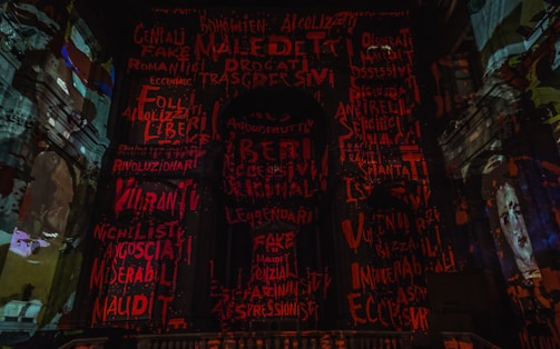 The image displays a dark, dramatic interior with walls covered in vibrant red and orange graffiti-style text. Architectural elements of what appears to be a historic building are visible. A portrait of a face is discernible on the right side.