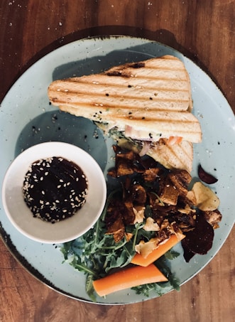 A toasted sandwich with grill marks is placed on a blue plate alongside a small bowl of sauce garnished with sesame seeds. Fresh greens, carrot sticks, and crispy vegetable chips surround the sandwich, creating a visually appealing arrangement.