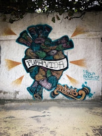 A colorful graffiti mural featuring a stylized heart with swirling patterns in various shades of teal, orange, purple, and black. Text on the heart reads 'PURA VIDA' in bold letters. Additional graffiti tags are present, including 'DRENAS' in an ornate script and 'ISLA SOUND!' in a blue stencil font. The mural is painted on a textured white wall with leaves hanging from the top edge.