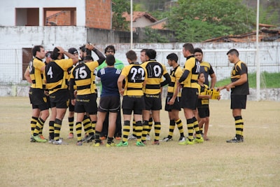 A group of rugby players are huddled together on a field wearing black and yellow uniforms. Two individuals in different outfits seem to be coaching or giving instructions. There is a child also in a similar uniform holding some equipment. The background includes a white concrete wall with some buildings and trees visible.