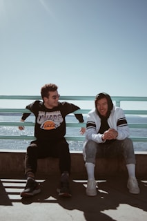 Two people are sitting on a seaside promenade with metal railings. One is wearing a black Los Angeles Lakers sweatshirt, sunglasses, and sneakers. The other person is in a white and black hoodie, rolled-up jeans, and white sneakers. They are sitting on a concrete ledge, enjoying the sunny day.