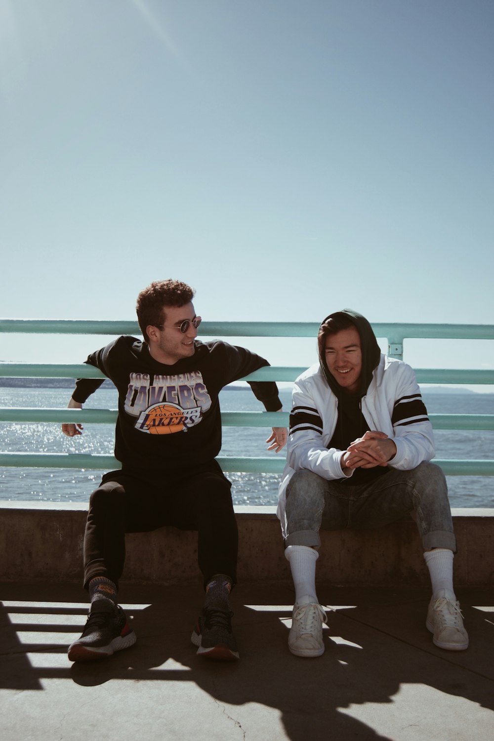 two smiling men sitting on fence near the body of water outdoor during daytime