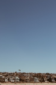A coastal residential area is visible with several modern houses lined up along a sandy beach. The sky is clear and blue, and there are a few palm trees scattered among the homes.