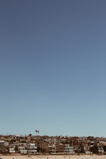 A coastal residential area is visible with several modern houses lined up along a sandy beach. The sky is clear and blue, and there are a few palm trees scattered among the homes.