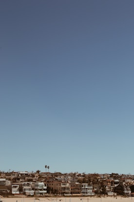 A coastal residential area is visible with several modern houses lined up along a sandy beach. The sky is clear and blue, and there are a few palm trees scattered among the homes.