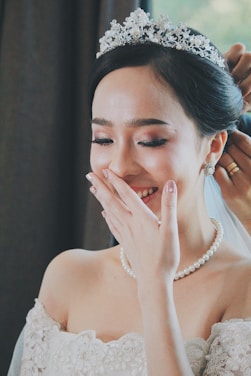 A bride is smiling joyfully with her hand covering part of her face, wearing an elegant white wedding dress, a pearl necklace, and a decorative tiara. Another person is adjusting her hair at the back.