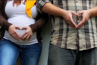 Two people are standing side by side with their hands forming heart shapes over a pregnant belly. The person on the left is wearing a brown sweater over a white top and blue jeans, and the person on the right is wearing a plaid shirt and jeans. A yellow scarf or accessory is visible around the neck of the person on the left.
