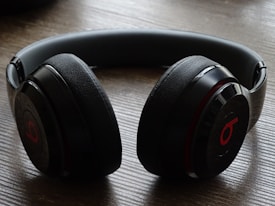 A pair of over-ear headphones with a sleek black design and a distinct red logo on the ear cups. The headphones are resting on a wooden surface, featuring a modern and stylish appearance.