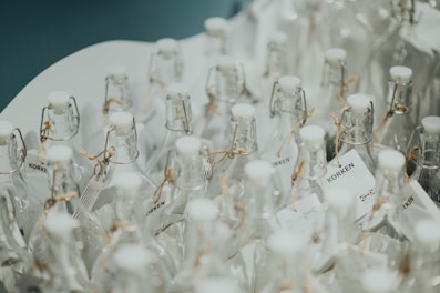 Numerous clear glass bottles with white caps arranged closely together, featuring cork-style closures and tags tied with twine. The surface reflects a clean and minimalistic aesthetic.