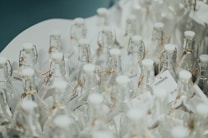 Numerous clear glass bottles with white caps arranged closely together, featuring cork-style closures and tags tied with twine. The surface reflects a clean and minimalistic aesthetic.