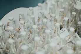 Numerous clear glass bottles with white caps arranged closely together, featuring cork-style closures and tags tied with twine. The surface reflects a clean and minimalistic aesthetic.