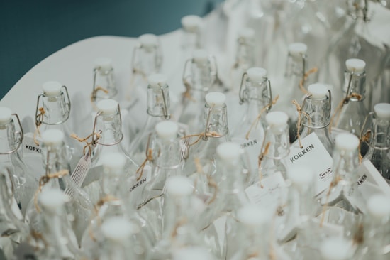 Numerous clear glass bottles with white caps arranged closely together, featuring cork-style closures and tags tied with twine. The surface reflects a clean and minimalistic aesthetic.