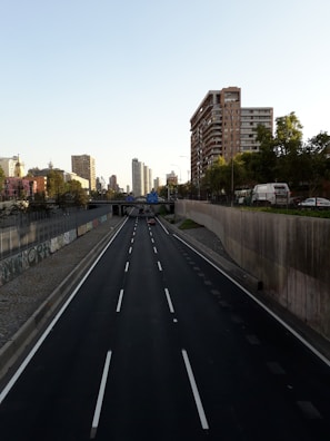 A multi-lane highway flanked by tall buildings under a clear sky. Several vehicles are visible on the road, including a red car and a truck. The sides of the highway feature concrete barriers and graffiti. Trees and urban structures surround the area, with a long view of the cityscape in the background.