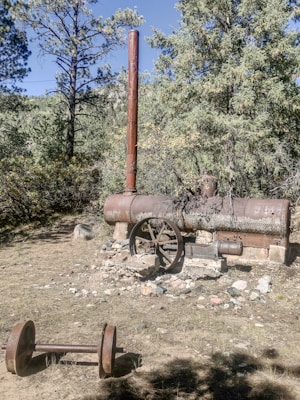 An old, rusted piece of industrial machinery is situated outdoors, surrounded by trees and natural vegetation. The machinery is cylindrical with a large, vertical exhaust pipe on top and a circular wheel on one side. It appears to be a steam boiler or similar equipment from the past. On the ground nearby, a rusty axle with two wheels lies in the dirt, partially covered by shadows.