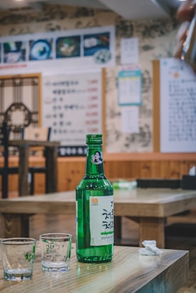 A green glass bottle with Korean text and an image of a person is placed on a wooden table. Next to the bottle are two small, empty shot glasses. In the background, there is a restaurant setting with Korean menus and decorations on the wall.