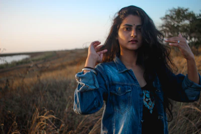 A model wearing a classic denim jacket and cowboy boots standing beside a wooden fence at sunset.