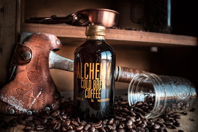 A brown glass bottle labeled 'Alchemy Cold Brew Coffee' is placed on a wooden surface surrounded by coffee beans. Next to the bottle is an overturned clear glass jar with more coffee beans scattered around. In the background, a rustic shelf holds a leather-handled axe and a metallic copper cup.
