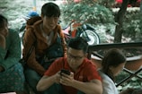A group of young people sitting together outdoors, two of them are engaged with their smartphones, while one looks towards another person, appearing to be in conversation. A motorcycle is visible in the background with lush green surroundings.