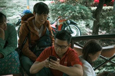 A group of young people sitting together outdoors, two of them are engaged with their smartphones, while one looks towards another person, appearing to be in conversation. A motorcycle is visible in the background with lush green surroundings.