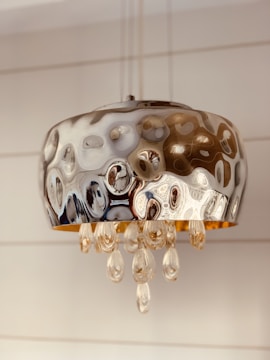 A modern metallic pendant light with a circular design, featuring a rippled surface and hanging crystal-like ornaments. The light fixture has an elegant and reflective quality, with a warm, ambient glow.