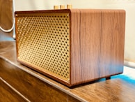 A wooden speaker with a lattice front design made of gold-colored material. It is placed on a wooden surface and has a retro, elegant appearance with smooth wood grain texture.