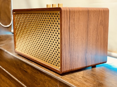 A wooden speaker with a lattice front design made of gold-colored material. It is placed on a wooden surface and has a retro, elegant appearance with smooth wood grain texture.