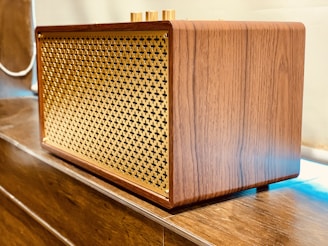 A wooden speaker with a lattice front design made of gold-colored material. It is placed on a wooden surface and has a retro, elegant appearance with smooth wood grain texture.