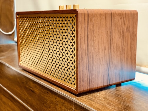 A wooden speaker with a lattice front design made of gold-colored material. It is placed on a wooden surface and has a retro, elegant appearance with smooth wood grain texture.