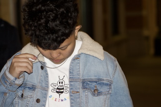 A person with short curly hair, wearing a denim jacket with a shearling collar, and holding a chain around their neck. The person is looking down, and their white shirt features a cartoon bee with the text 'Breezway' beneath it.