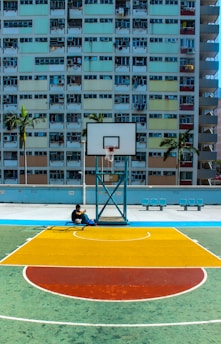 man sitting below basketball hoop near the building during daytime