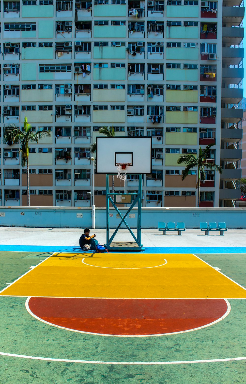 man sitting below basketball hoop near the building during daytime
