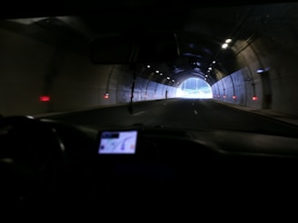A view from inside a car driving through a tunnel, with visible dashboard and GPS. The tunnel is dimly lit, with a bright exit visible ahead. Red indicators on the tunnel walls provide guidance.