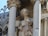 A stone sculpture of an angel with curly hair and a gentle smile, positioned between two ornate columns. The angel holds a draped garment and features detailed wings. The background includes gothic architecture elements and decorative carvings.