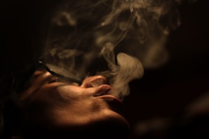 A close-up of a person exhaling smoke, with soft lighting creating a dramatic and moody atmosphere. The smoke is wispy and curling around the face, providing an ethereal effect.
