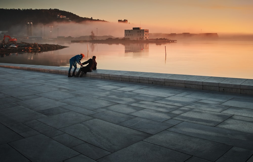 two person sitting on concrete floor near body of water during golden hour
