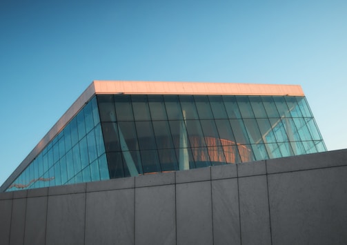 A modern architectural structure featuring large glass walls and a flat roof with a soft pink hue from the sunlight. The front glass facade has a grid-like pattern, and the building rests on a solid gray foundation.